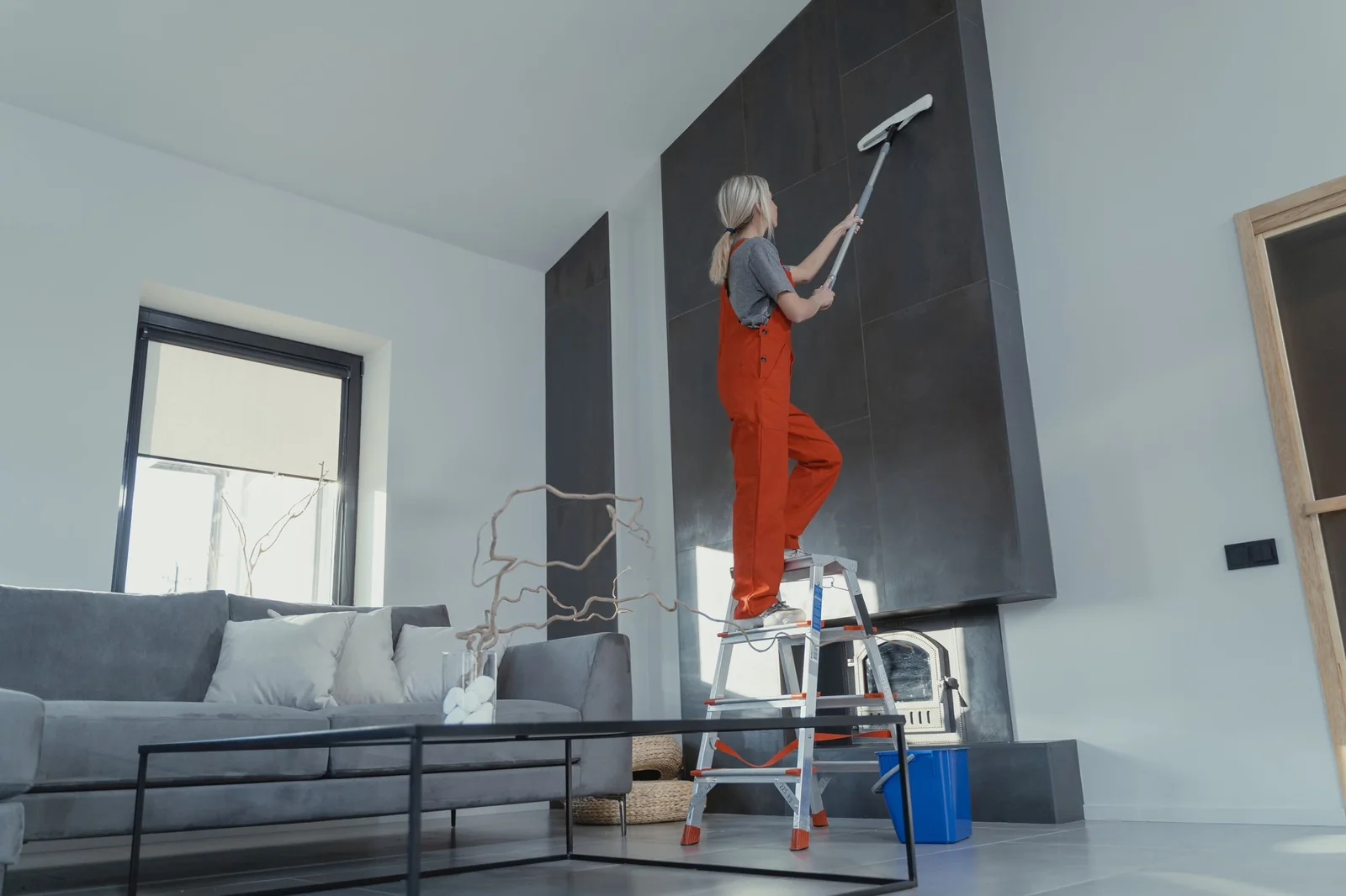 Cleaner on a step ladder dusting high surfaces in a bright interior